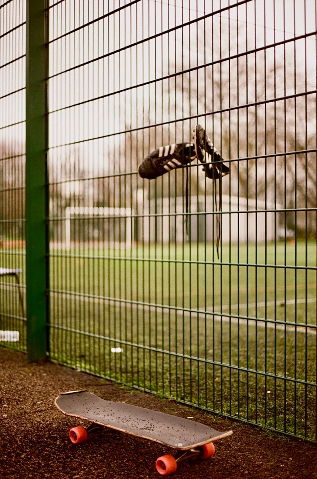 Football boots left in a wire fence surrounding a deserted football pitch, skateboard on the ground below.