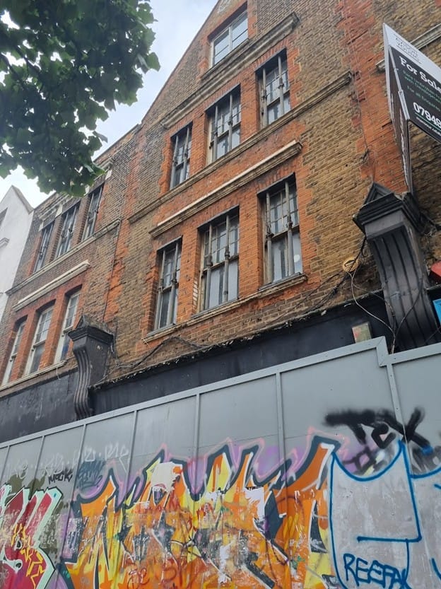 Victorian buildings seen above boarding with graffitti.  Upper floors were vacant before 2019 plans.