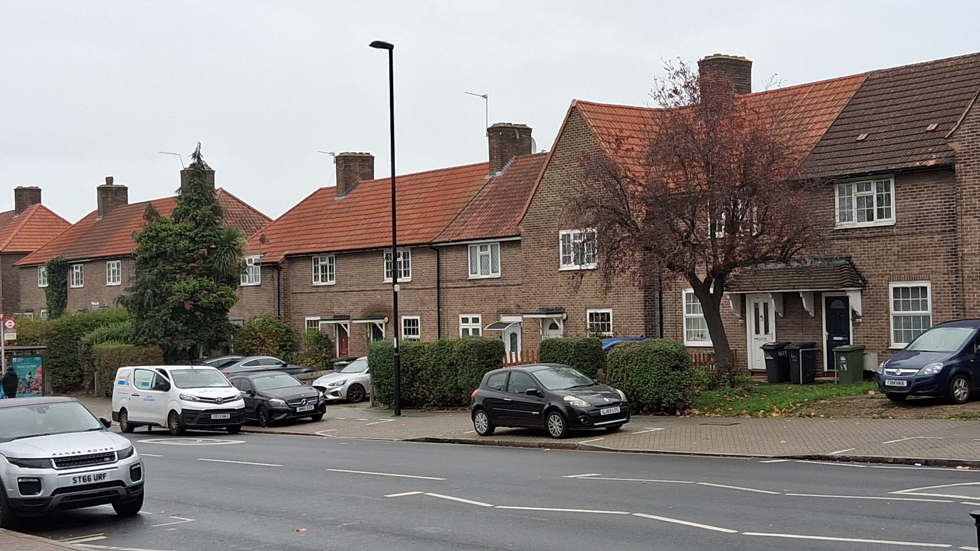 Row of red brick, terraced houses, small front gardens, traffic on busy road.