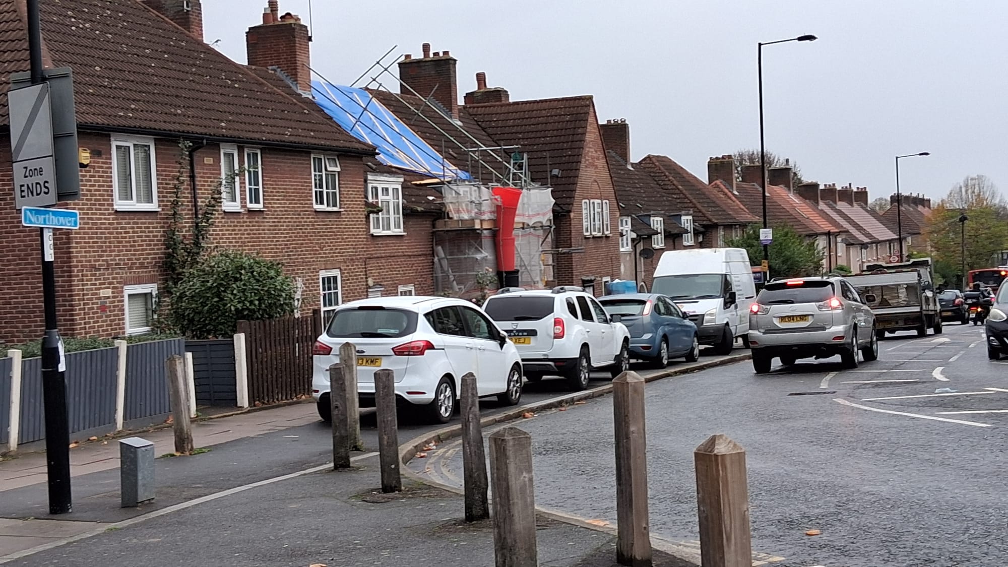 Row of red brick, terraced houses, with cars and van parked on pavement, traffic on road.