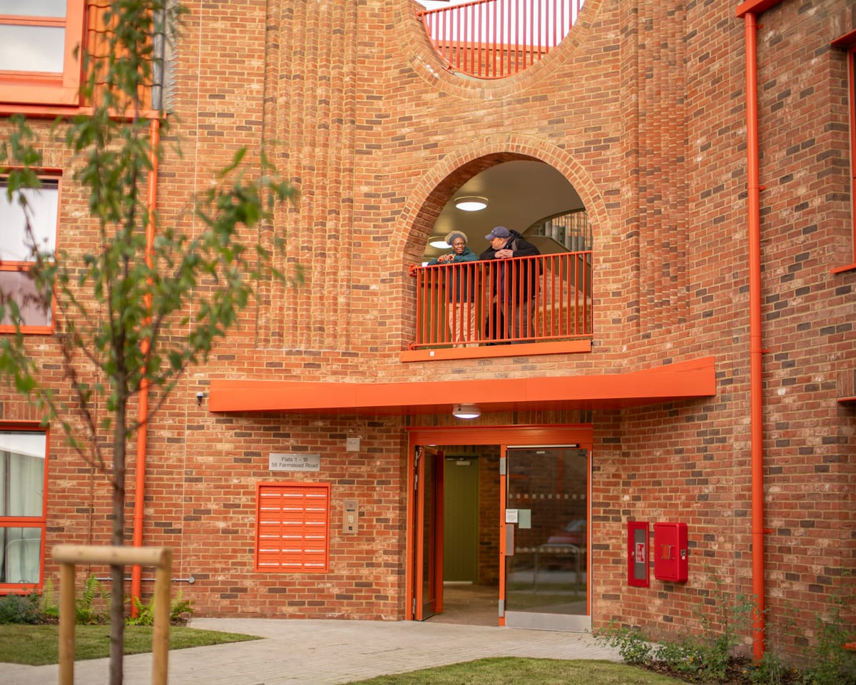 Two residents on balcony above entrance Farmstead road, 3 story building, red brick facade with red balcony with arch