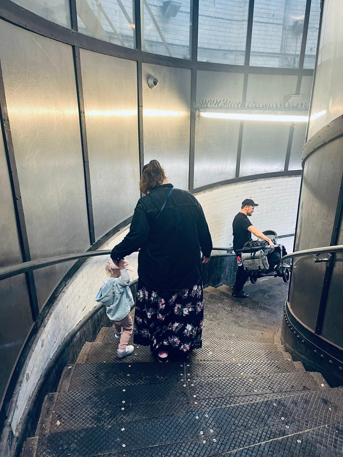 A young family navigate the stairs, man carries buggy while very young child walks down steps with mother