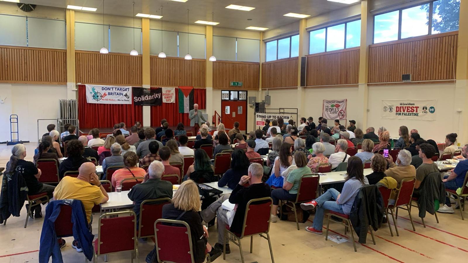 Crowded hall with around 40 people listening to a speaker. Banners on wall including Divest for Palestine