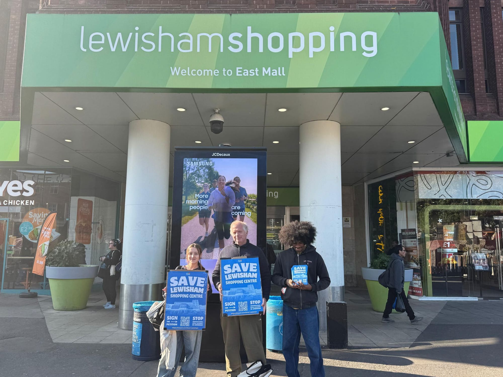 3 campaigners outside Lewisham Shopping Centre with placards "Save Lewisham Shopping Centre".