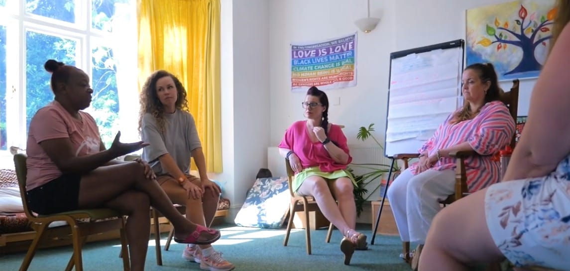 Scene of 5 participants and co-researchers sitting in workshop in sunny room, one woman talking.