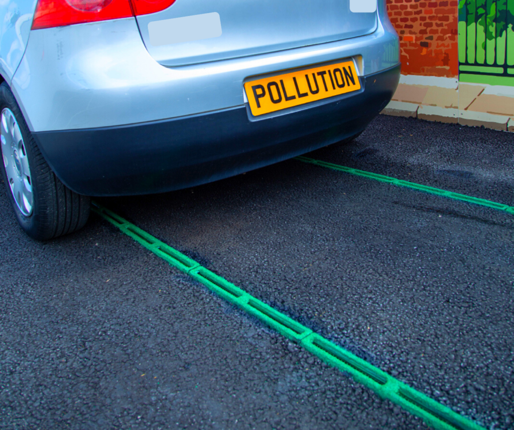 Car with numberplate "Pollution" parked over narrow vents in the road, with green edging. Painted backdrop of brick wall.