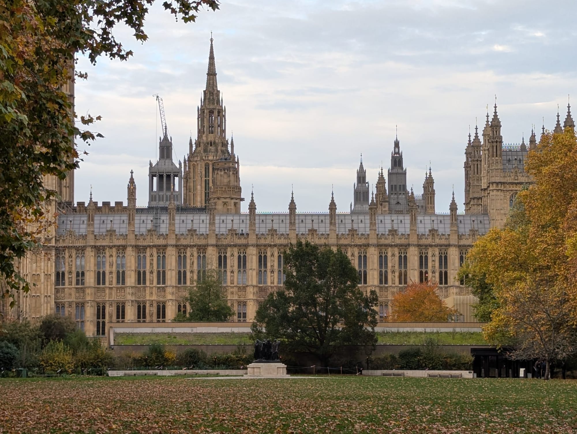 Palace of Westminster, seen from Victoria Tower Gardens South. Framed by autumn trees with leaves on grass. 