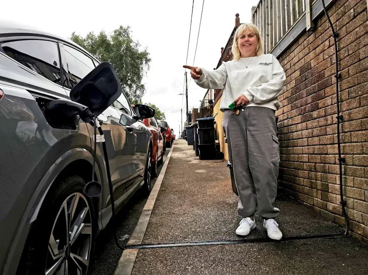 First install in Wales, smiling woman pointing to charging car, she is standing over cable in gulley in pavement.
