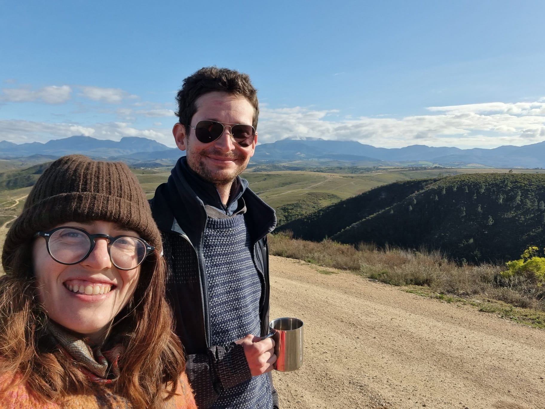 Hosts Tommie and Jack in winter jackets on a hillside, smiling for selfie, rolling hills behind.