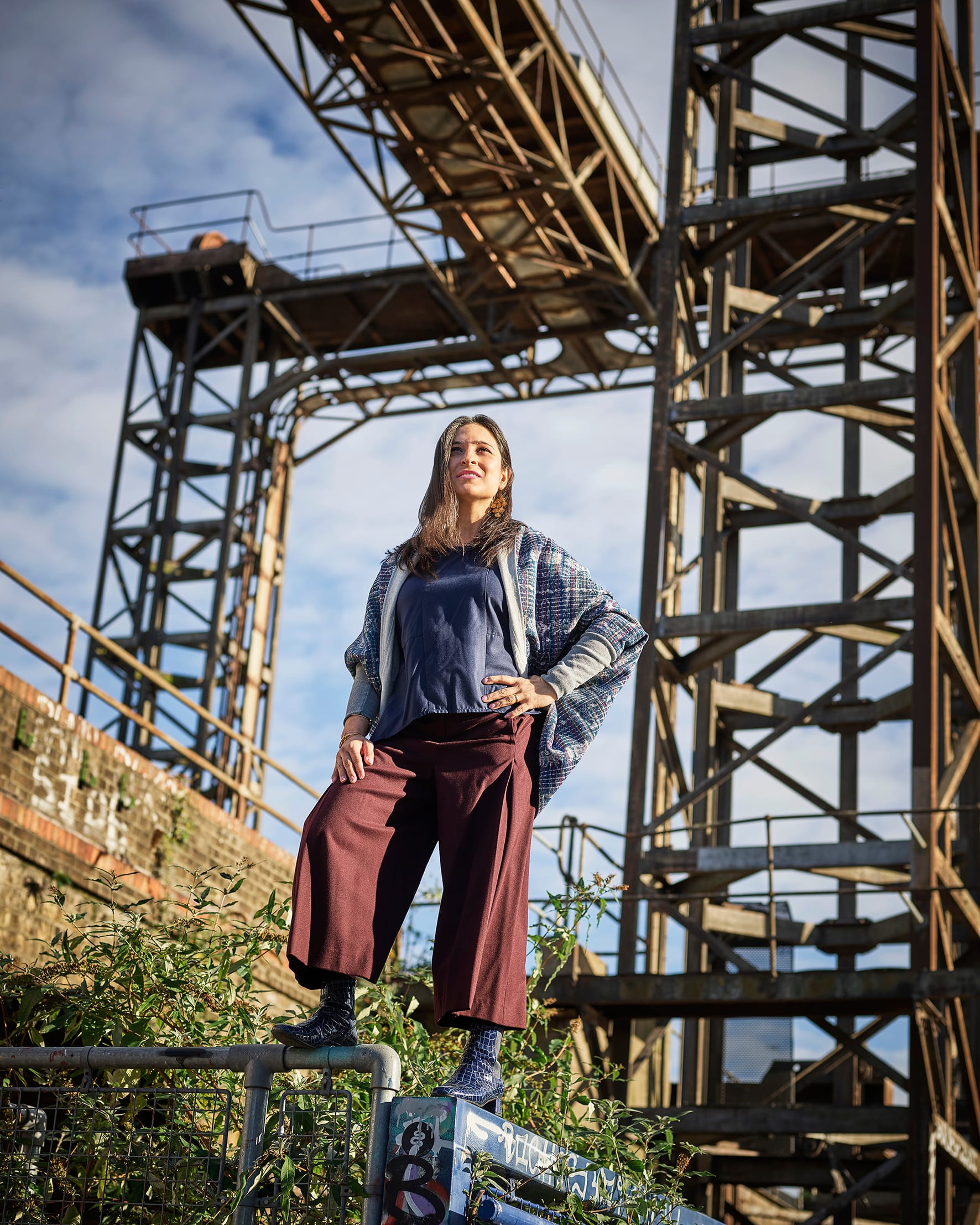Rupa Ganguli in wide leg trousers and boots, perched on a wall, budlea and the creek bridge behind her. 