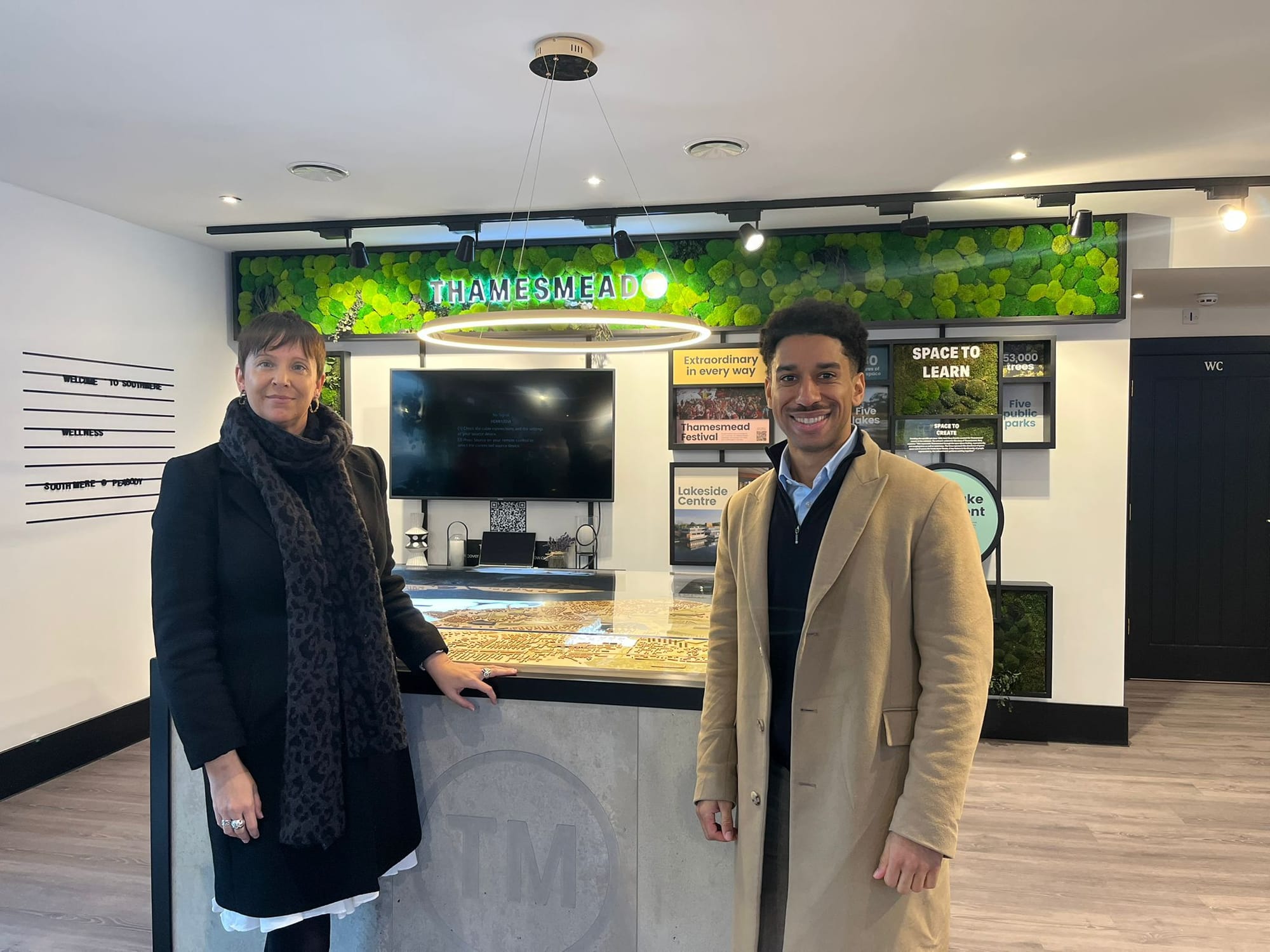 London Assembly Members Elly Baker and James Small-Edwards indoors but wearing coats, smiling.