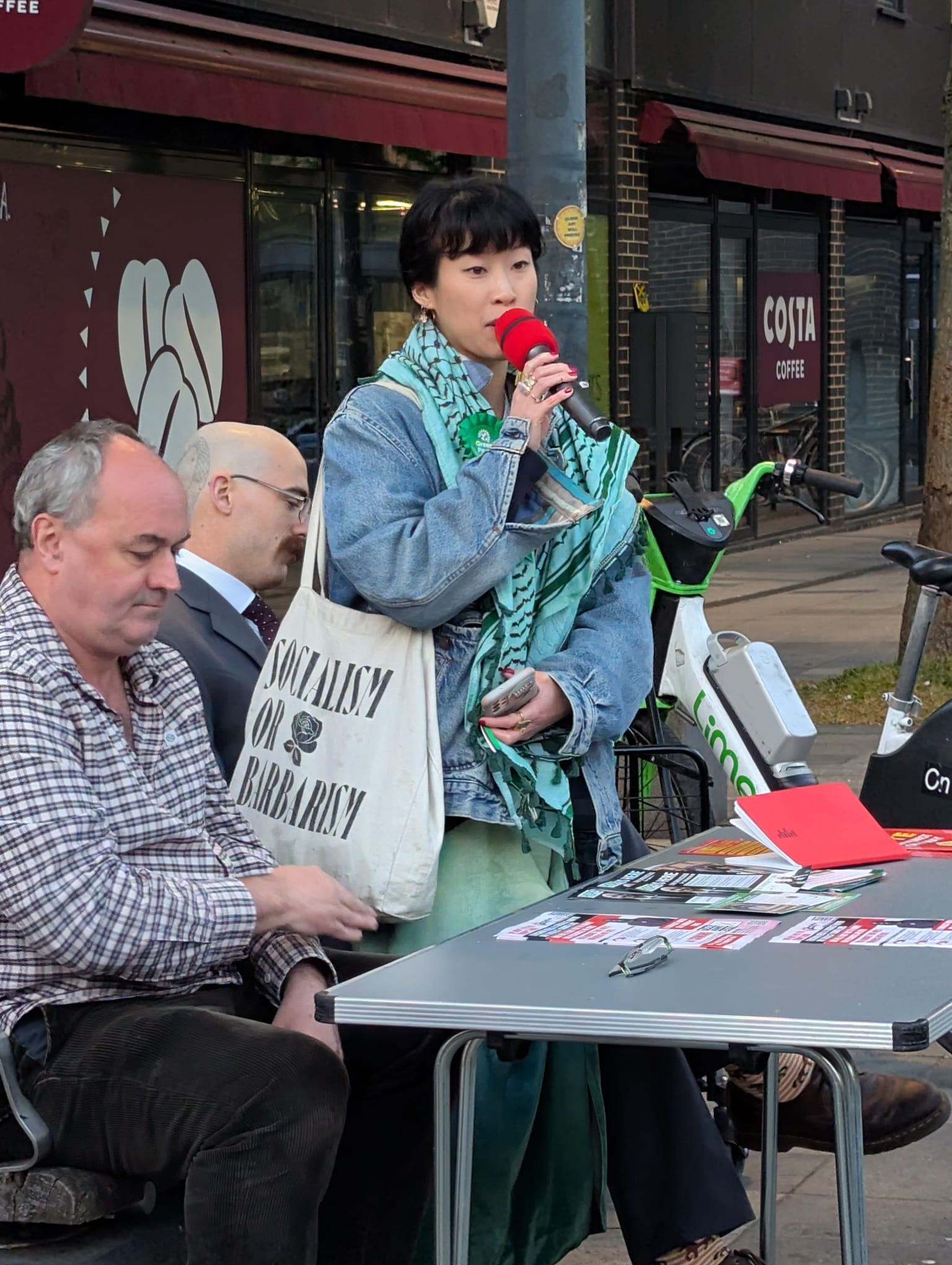 Young woman in denim jacket, green-tinged keffiyeh speaks to mic.  Bag over shoulder with slogan "Socialism or Barbarism". 