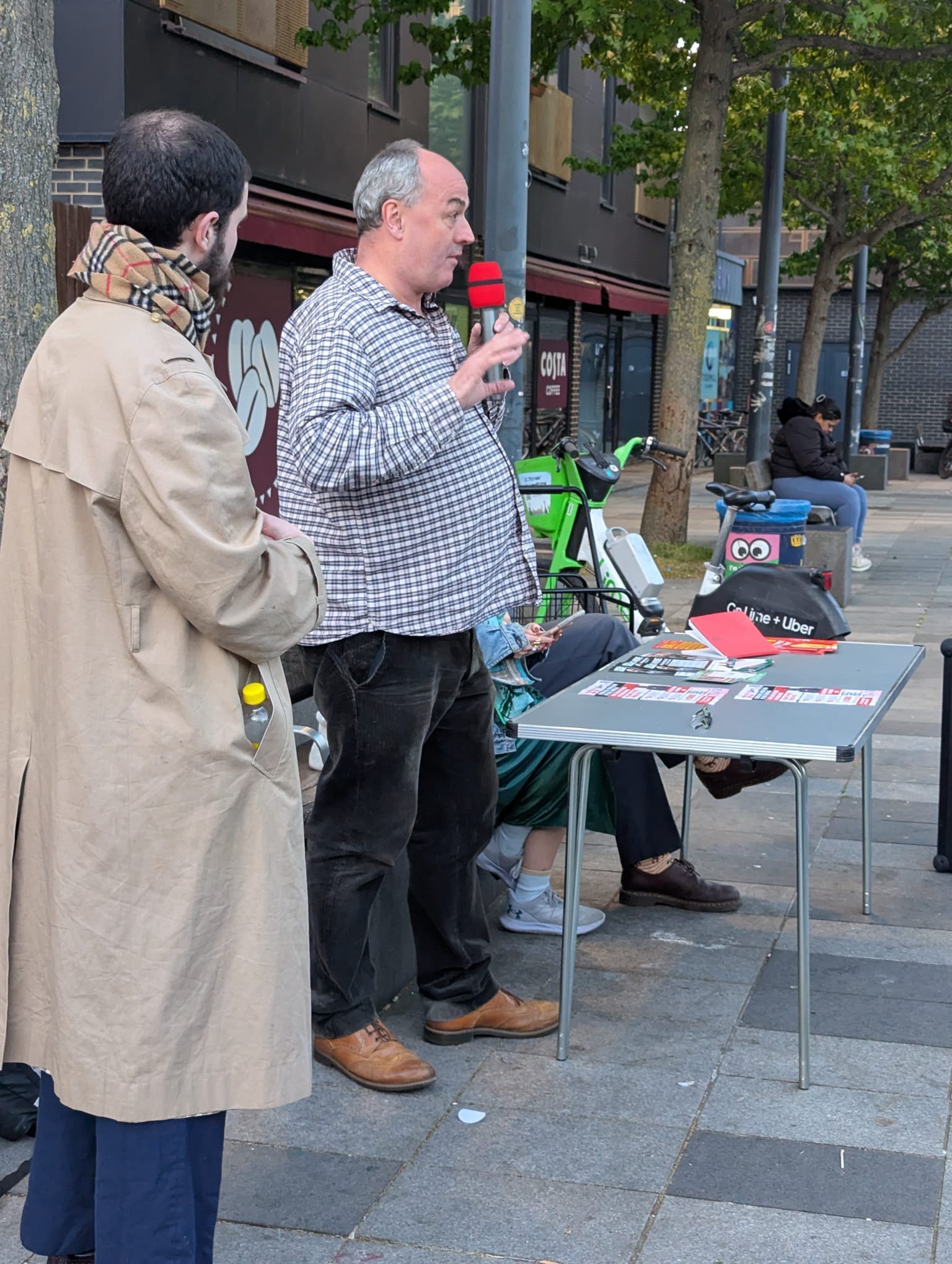 Man in checked shirt speaking into mic at outdoor event on a square.