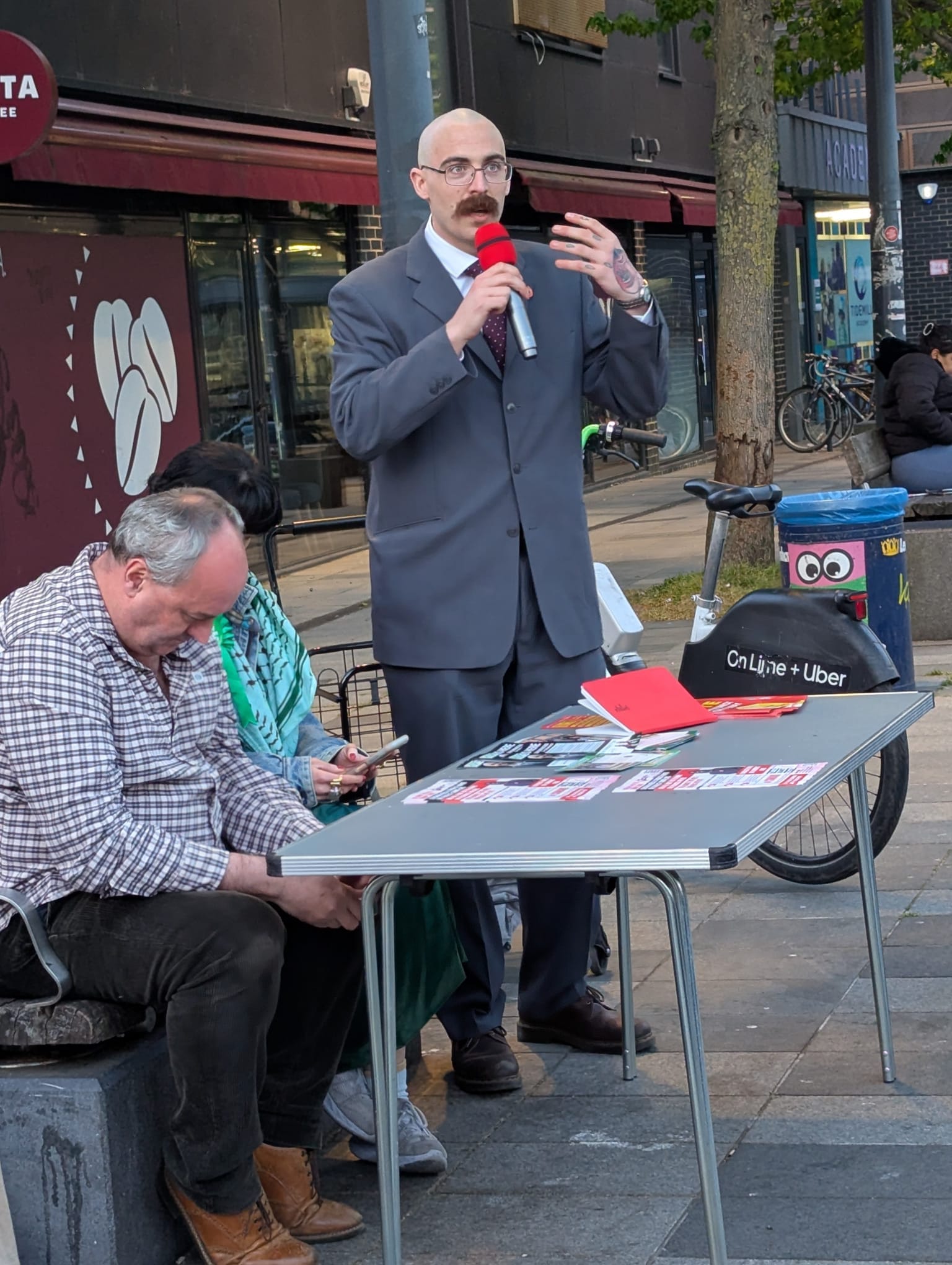 Bald man in old-fashioned grey suit and tie, with glasses, moustache and tatoo on hand speaking to mic