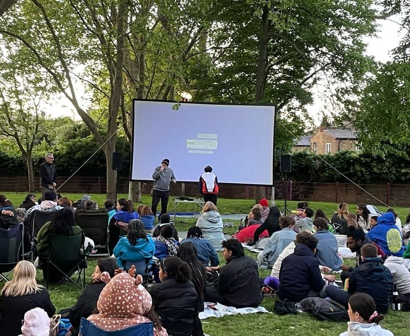 Showing of outdoor film in Eckington Gardens, people sitting on the grass with a large screen set up with guy ropes. 