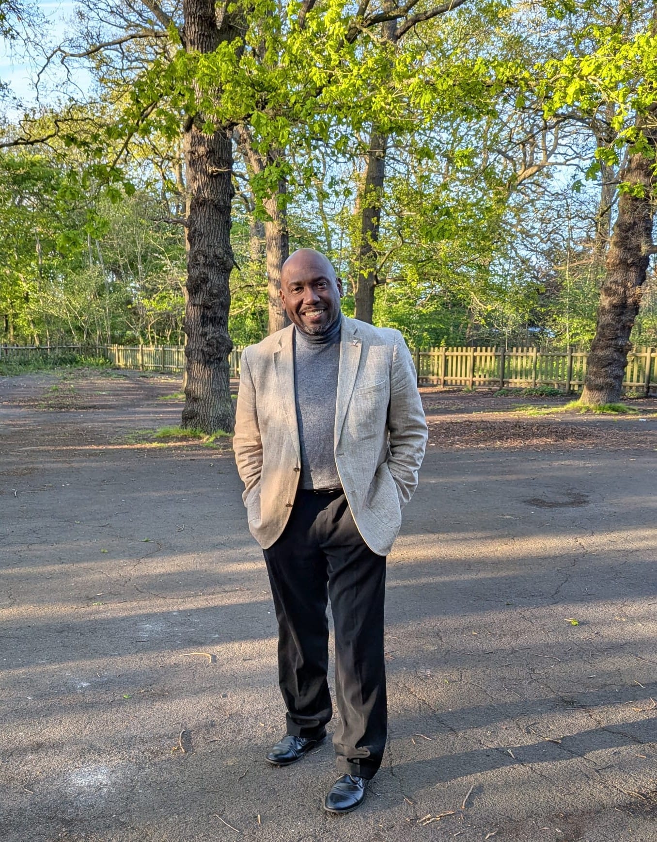 Man in casual jacket, smiling, hands in pockets with trees and wooden fence behind.