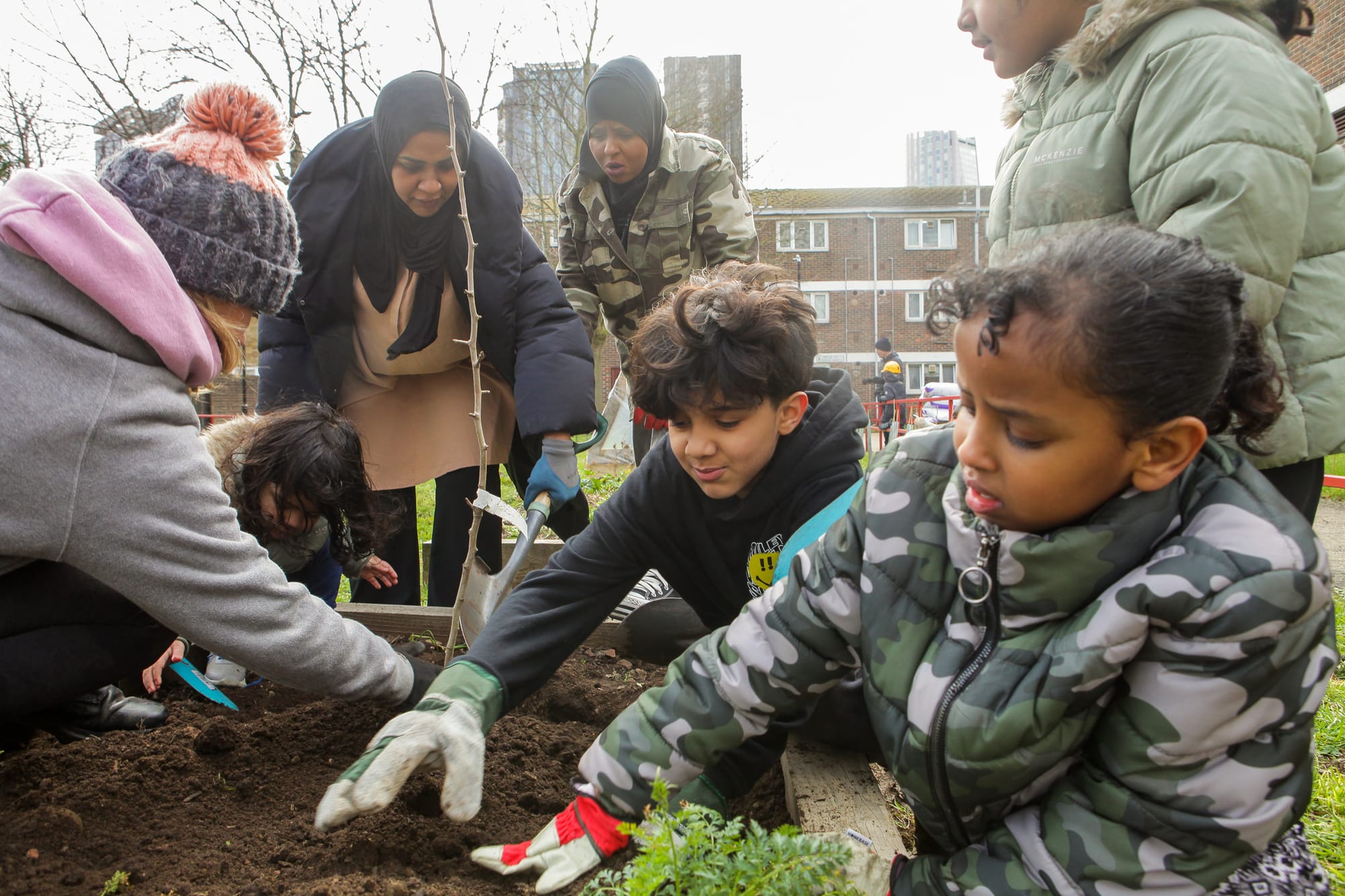 Children planting a young tree in a raised bed, estates visible behind.
