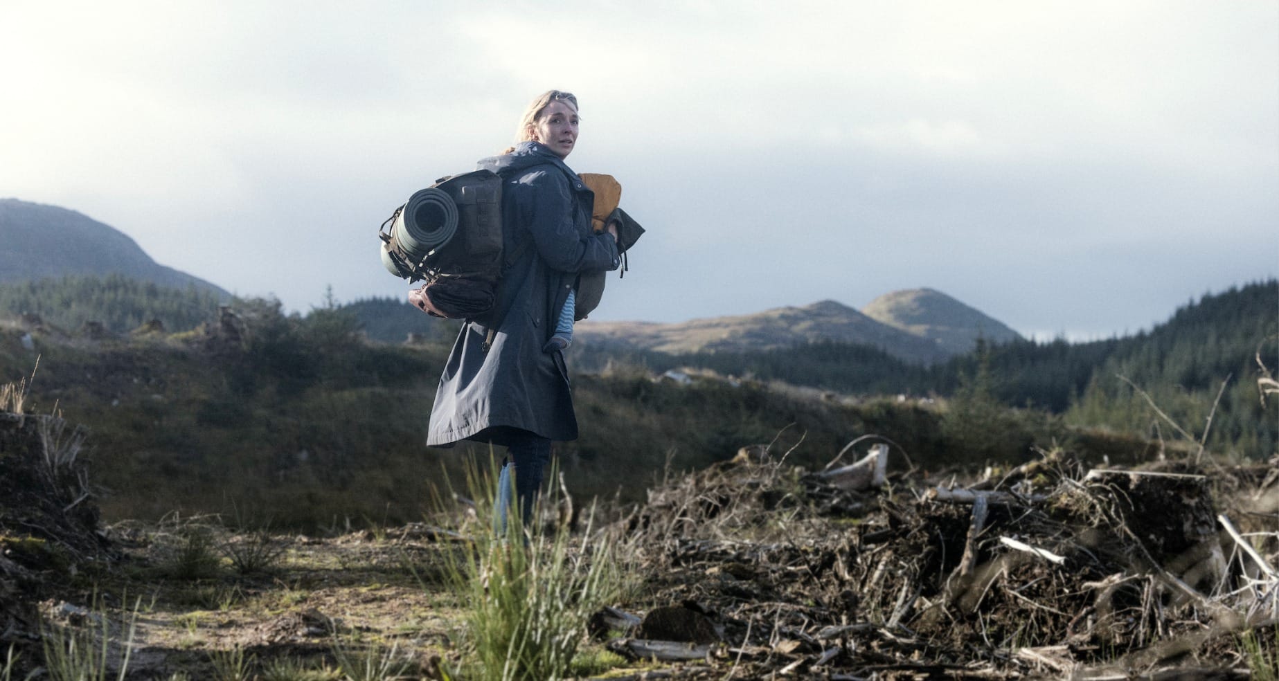 Woman with backpack and sleeping mat on her back, carrying young baby in a sling, in a remote and barren, hilly landscape.