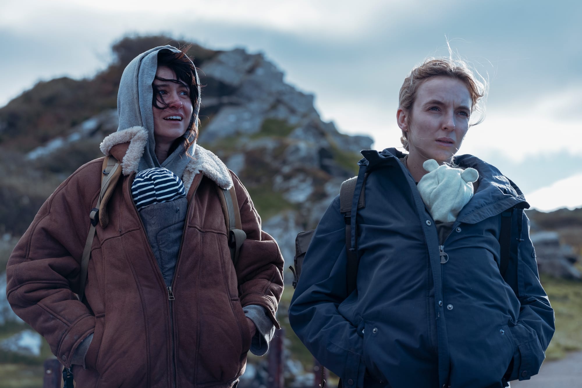 Two women standing side by side in a remote and barren landscape, one speaking and smiling. Each carries a baby in a sling with their jackets buttoned over it.