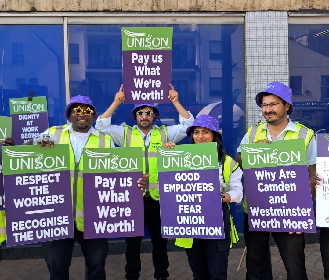 4 smiling parking officers in hi-vis jackets and purple bucket hats, in picket line outside the town hall with placards: "Respect the Workers, Recognise the Union".