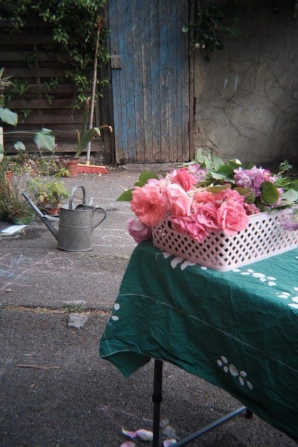 Corner of table with basket of pink roses on it, watering can in the background with old wooden door.