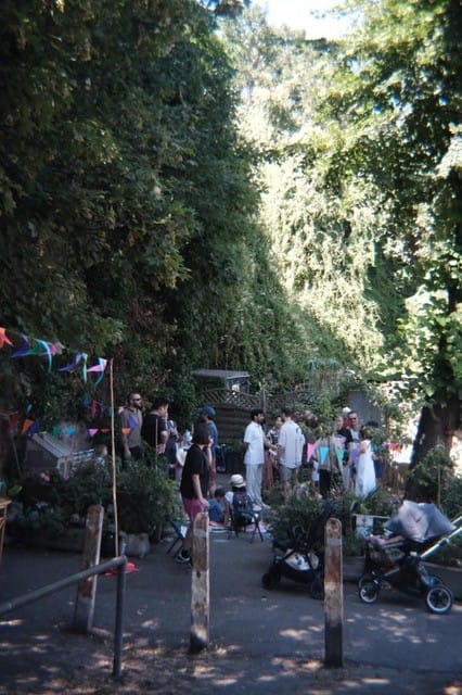 Gathering of people chatting in shade of large trees, some children and prams.