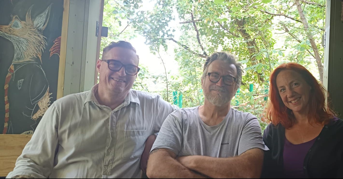 Fourth Reserve trustee Tommy Radclyffe-Thomas with co-founders Nick MacGuinness and Anna-Maria Cahalane sitting in hut, smiling, trees of reserve visible through window..