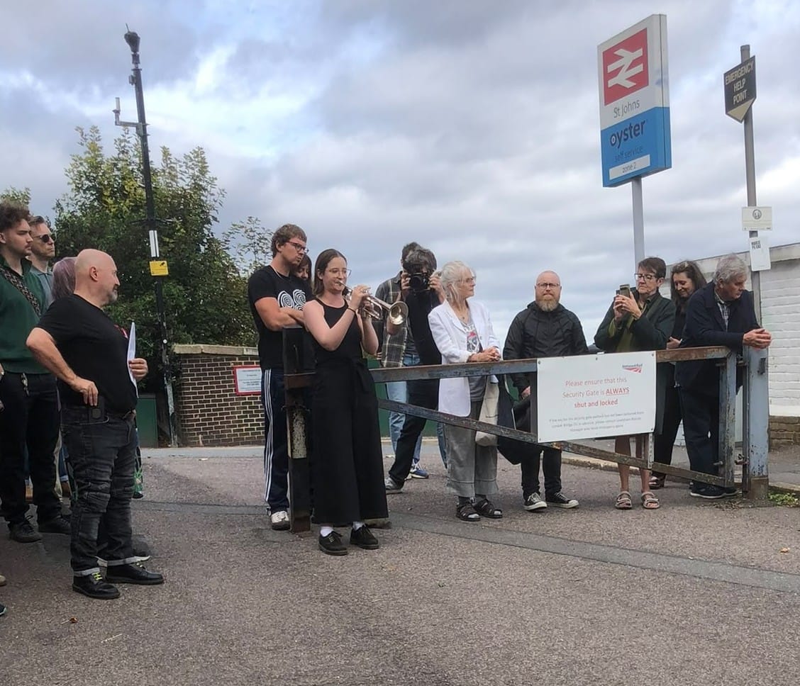 Young woman in black dress and trainers playing trumpet on the railway bridge, others watching.