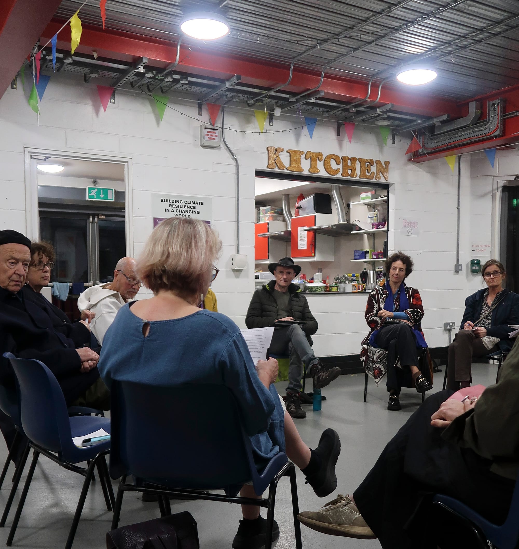 Participants sitting on plastic chairs in a circle, listening intently as a woman reads something out.