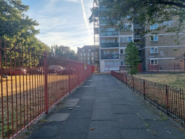 Iron railings on both sides of the footpath in the estate, dried grass on either side behind the railings. flats behind.