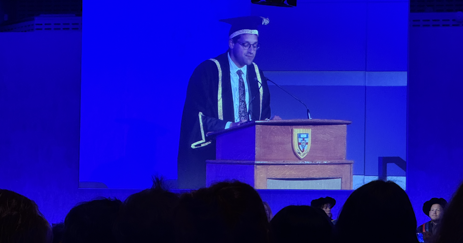 An academic in cap and gown at a lectern projected onto a blue screen, dark silhouettes of heads of students in auditorium watching.