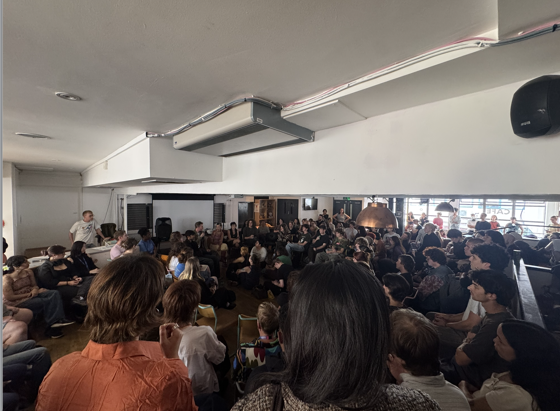 Crowded meeting in large hall or refectory, students listening to a speaker.
