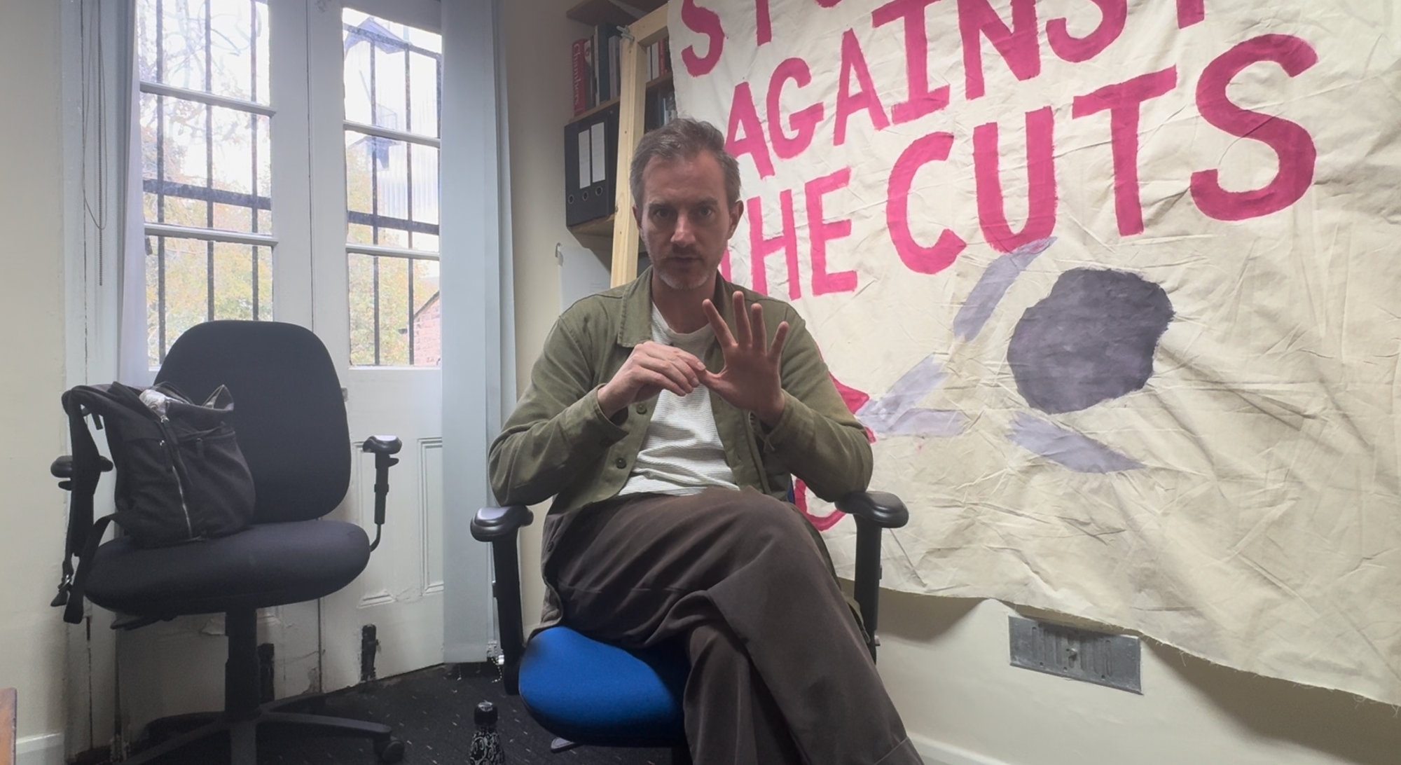 Man sitting on an office chair gesturing with his hands. Banner on wall with "Students against the cuts" sewn on in large pink letters.