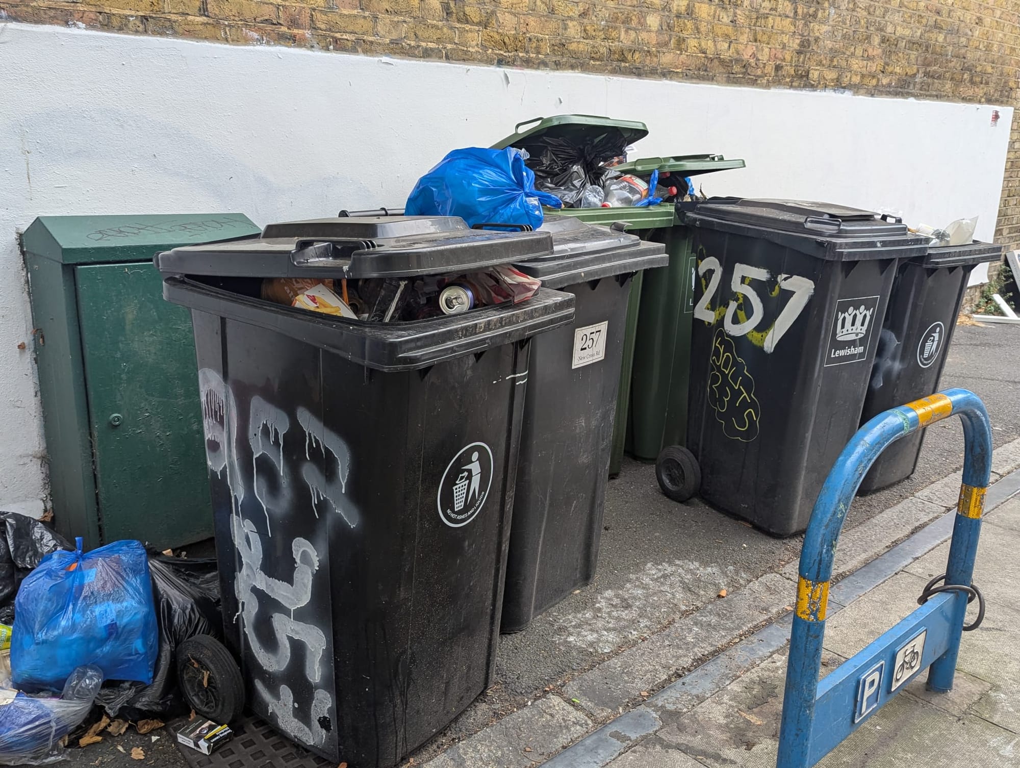 Overflowing black wheelie bins, with single overflowing recycling bin visible behind. Rubbish bags around bins on pavement.