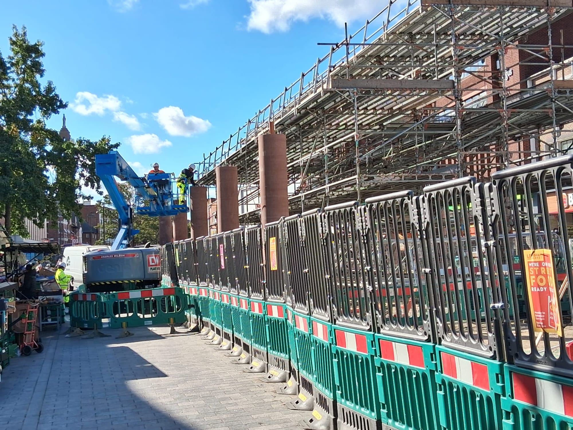 Construction work on Lewisham High Street, railings, digger, with worker in high vis in cherry picker, and scaffolding.