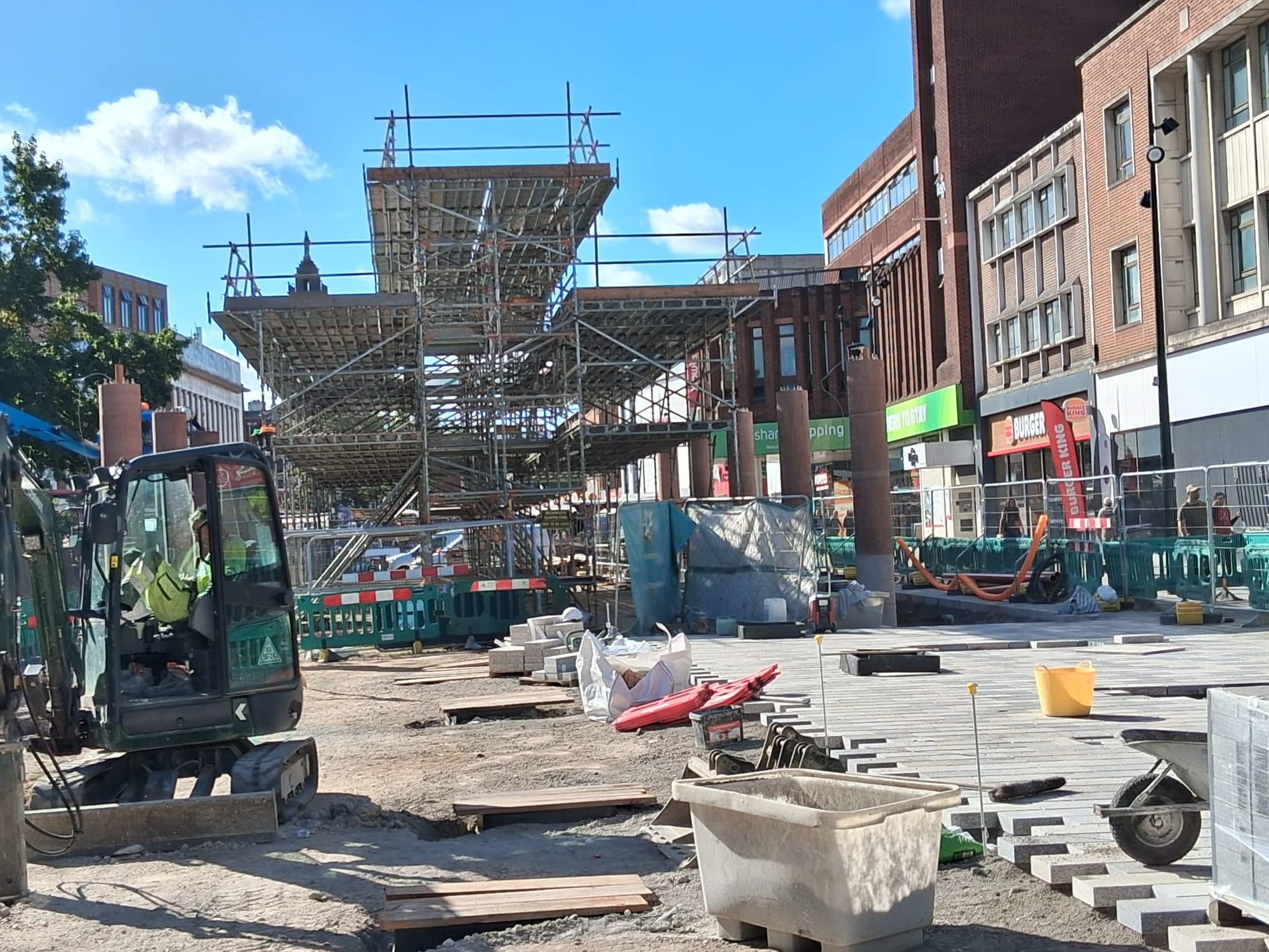 Construction work on Lewisham High Street, railings, worker in cab of digger, and scaffolding