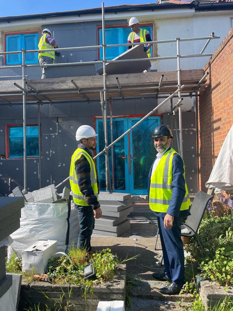 House with scaffolding on back wall, insulation blocks piled up in garden.  4 workers in picture in high vis and hard hats, 2 of them on the scaffolding, one lifting an insulation board.