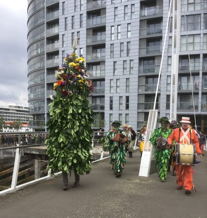 Parade of green man in headdress covered in leaves, drummers and accordion players crosses Greenwich Reach swing bridge  