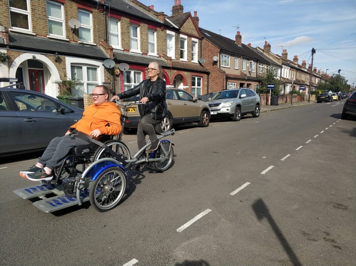 A disabled person in a wheelchair being driven on the road in a cycle-powered wheelchair carrier. 