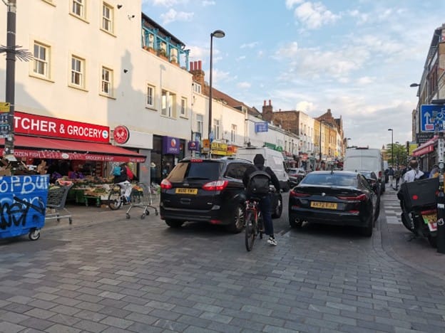 SUV negotiating around parked cars on Deptford High street while cyclist waits to pass.