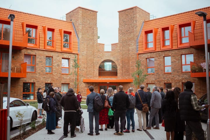 Small crowd at event. 3 story blocks joined at entrance, red brick facade, orange/red tiles, window frames, balconies