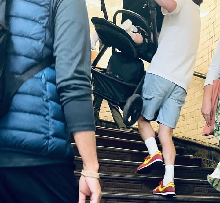 Man carries a buggy containing a small baby up the spiral stairs, other pedestrians seen.