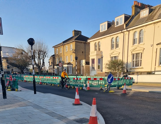 Newly widened pavement and new zebra crossing (under wraps) at Harefield Road and road works to widen pavement on west side of Brockley Road, cyclist navigating the single lane. 