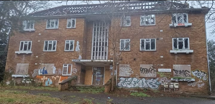 Large brick residential building, boarded up windows at ground level, smashed windows, graffiti, roof destroyed by fire