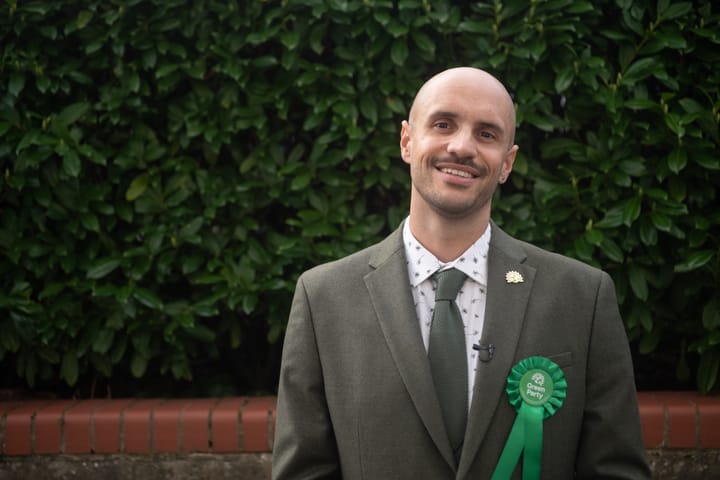 Liam Shrivastava smiling in suit with green rosette, green leaves behind. 