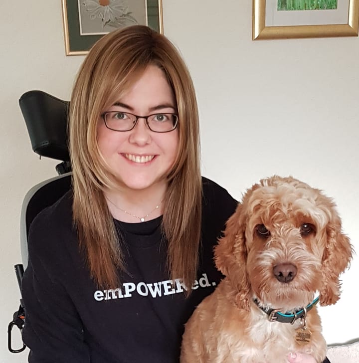 Woman sitting in wheelchair smiling with small cockadoodle dog sitting on her lap, dog staring at camera.