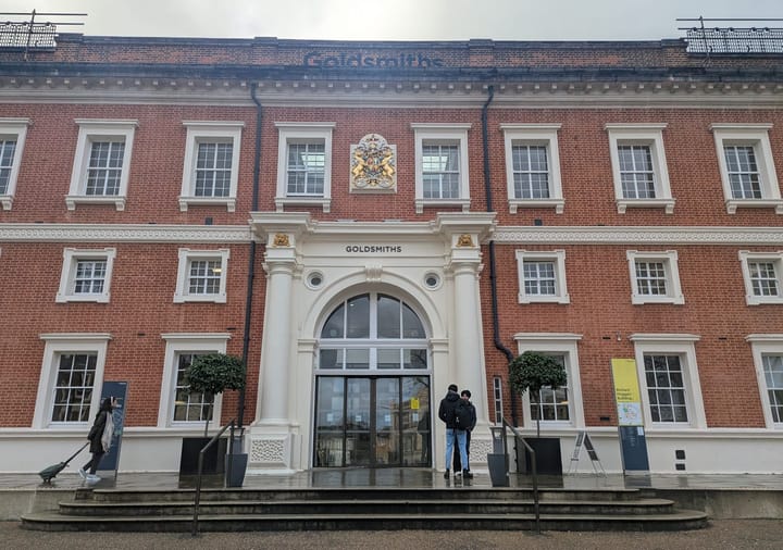 Goldsmiths university main entrance, red brick building, two students standing outside. Rain on steps.