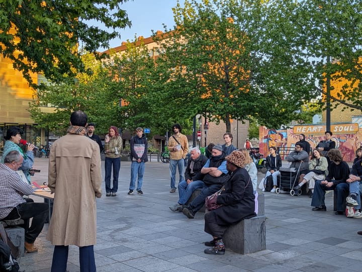 People standing in the square and sitting on benches listening to speakers in early evening sun at Deptford hustings. g