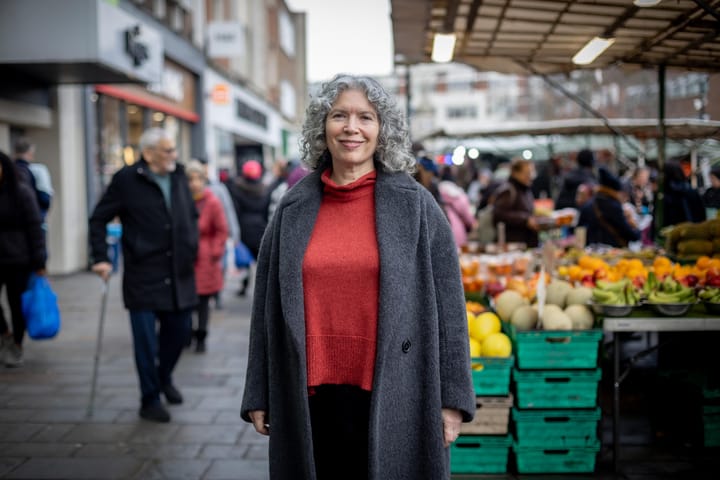 Amanda De Rky standing in grey coat, red jumper at the fruit and vegetable market outside Lewisham shopping centre.