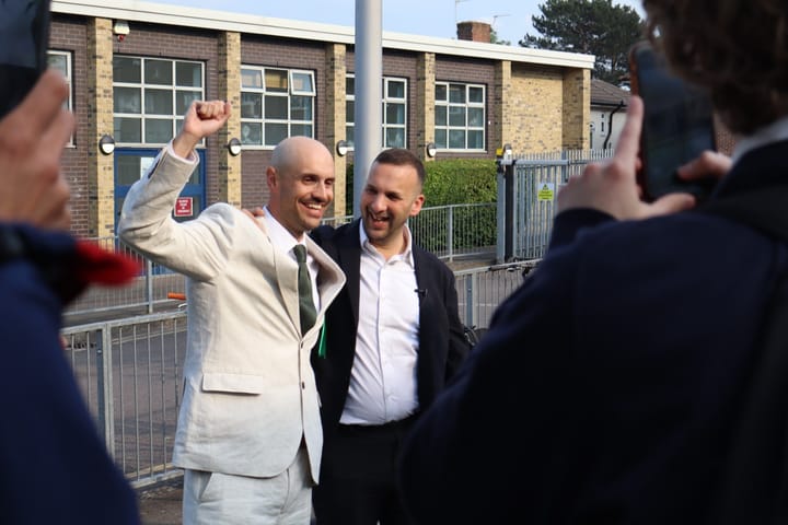 Liam Shrivastava grinning with raised fist, and Zack Polanski laughing outside the school.